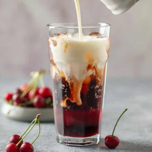 Cream being poured from a small pitcher into a tall glass of dark cherry cola over ice, creating swirling white ribbons through the drink, with fresh red cherries scattered around the glass.