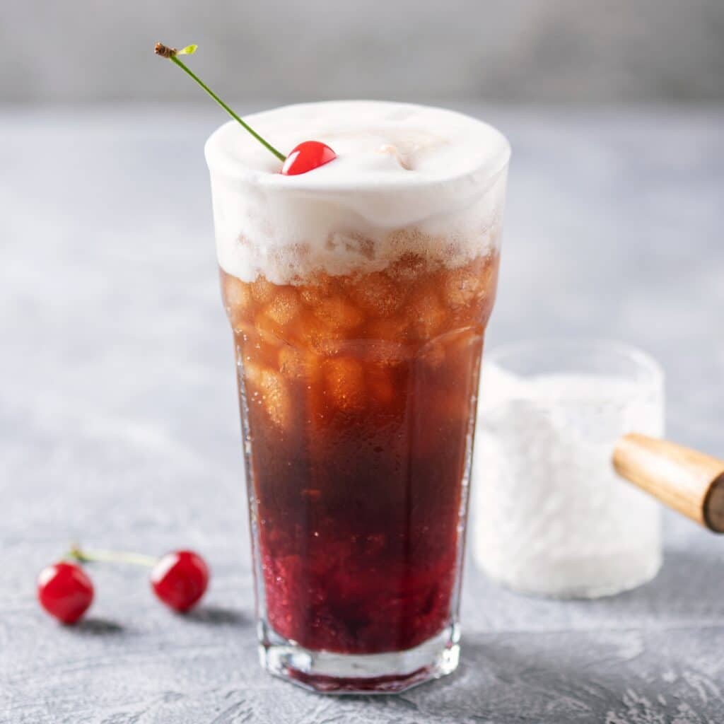 A tall glass of Dirty Dr Pepper topped with a thick dome of cream foam and a fresh cherry garnish, with more cherries and a small container of whipped foam beside it.