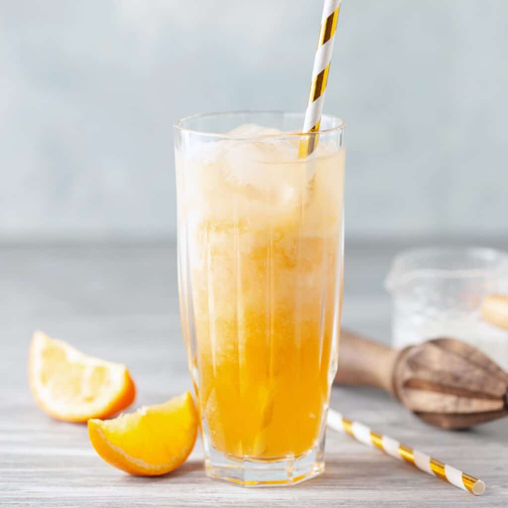 A tall ribbed glass of orange cream dirty soda with vibrant orange soda layered under swirled cream and a gold striped straw, with fresh orange slices and a wooden citrus juicer alongside.