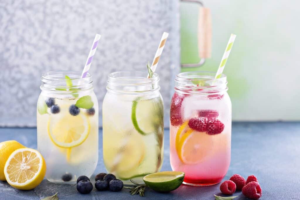 Three mason jar glasses with colorful striped paper straws contain fruit-infused water — lemon and blueberry, lime and rosemary, and pink raspberry lemon — with a vintage green cooler visible in the background.