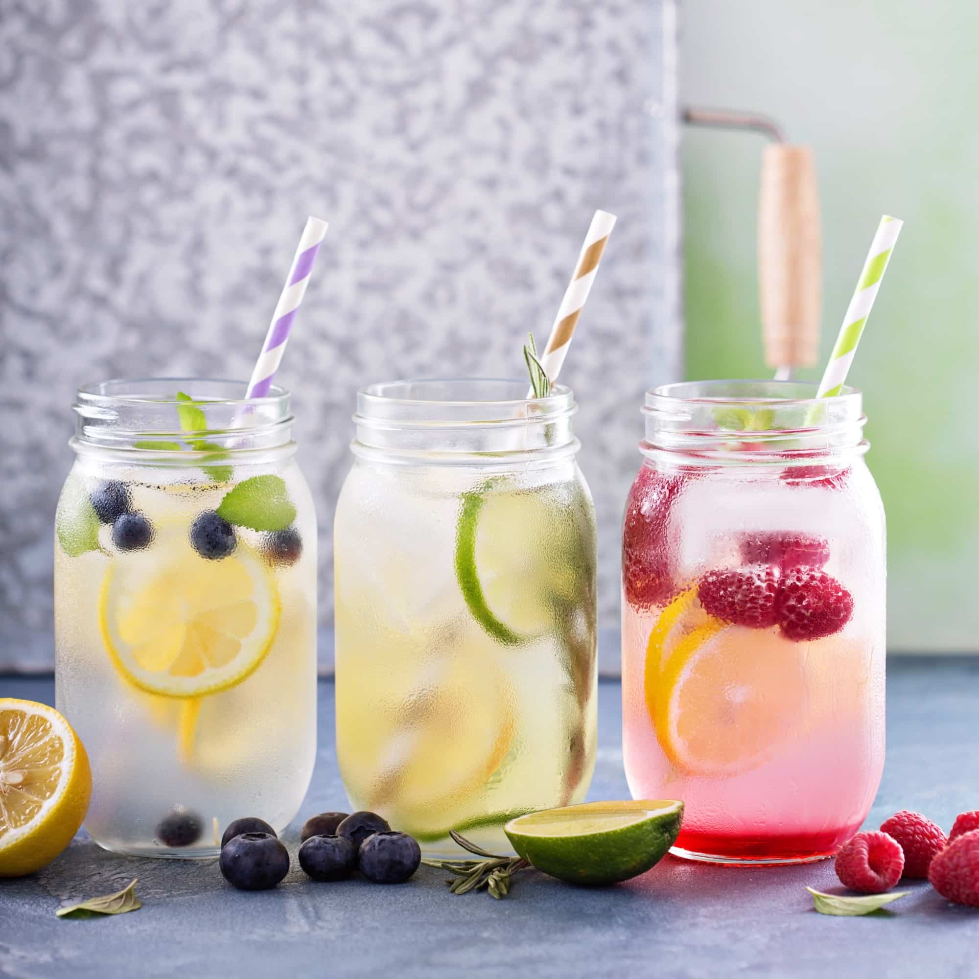 Three mason jar glasses with colorful striped paper straws contain fruit-infused water ā lemon and blueberry, lime and rosemary, and pink raspberry lemon ā with fresh fruit scattered on a blue-gray stone surface.