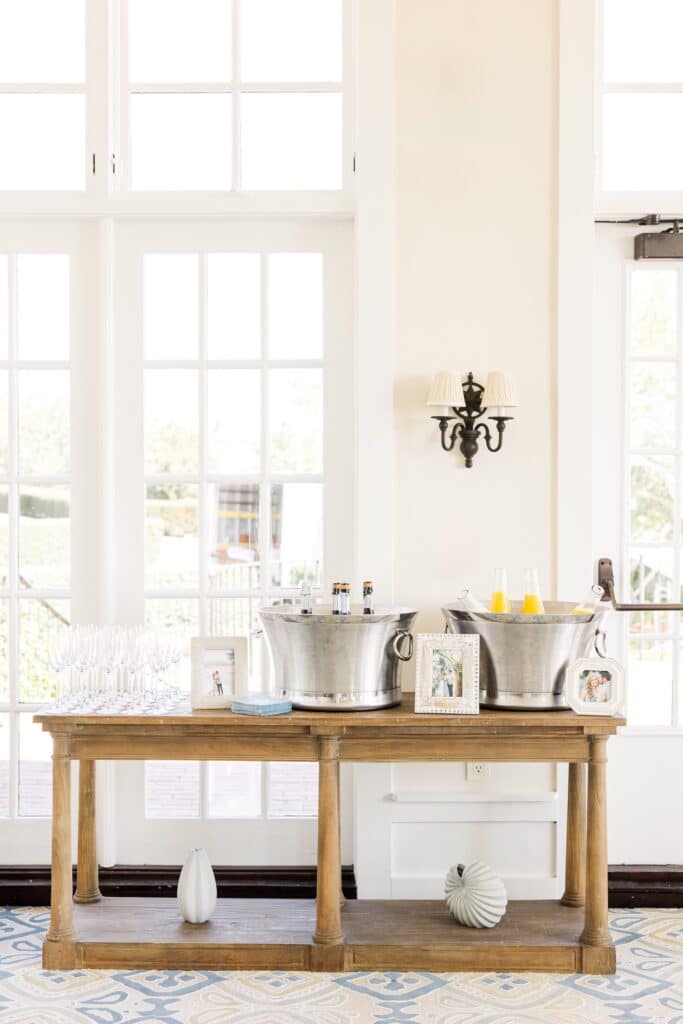Indoor mimosa station with two large silver galvanized tubs holding champagne bottles and orange juice on a wooden console table, champagne flutes lined up on the left and small framed photos displayed between the tubs.