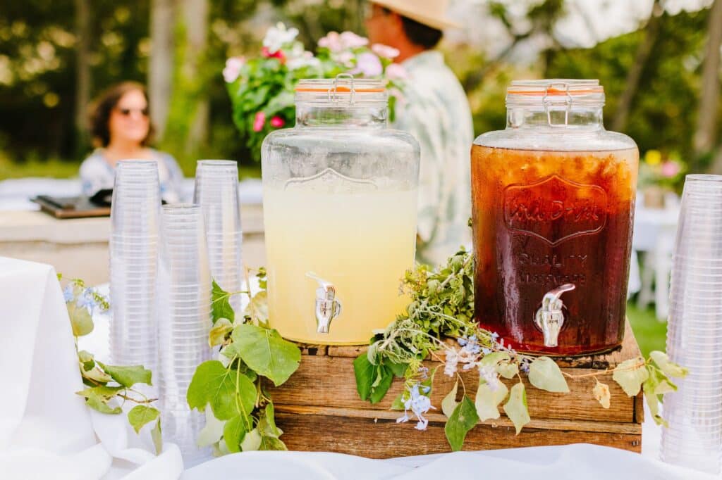Two large glass beverage dispensers filled with lemonade and iced tea sit on a wooden crate surrounded by a green floral garland, with stacked plastic cups on each side at an outdoor garden party.