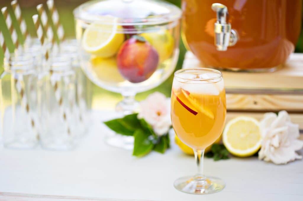 A stemmed glass filled with amber peach iced tea garnished with a peach slice sits at an outdoor drink station alongside glass bottles with gold striped straws, a bowl of peaches and lemons, and a large beverage dispenser.