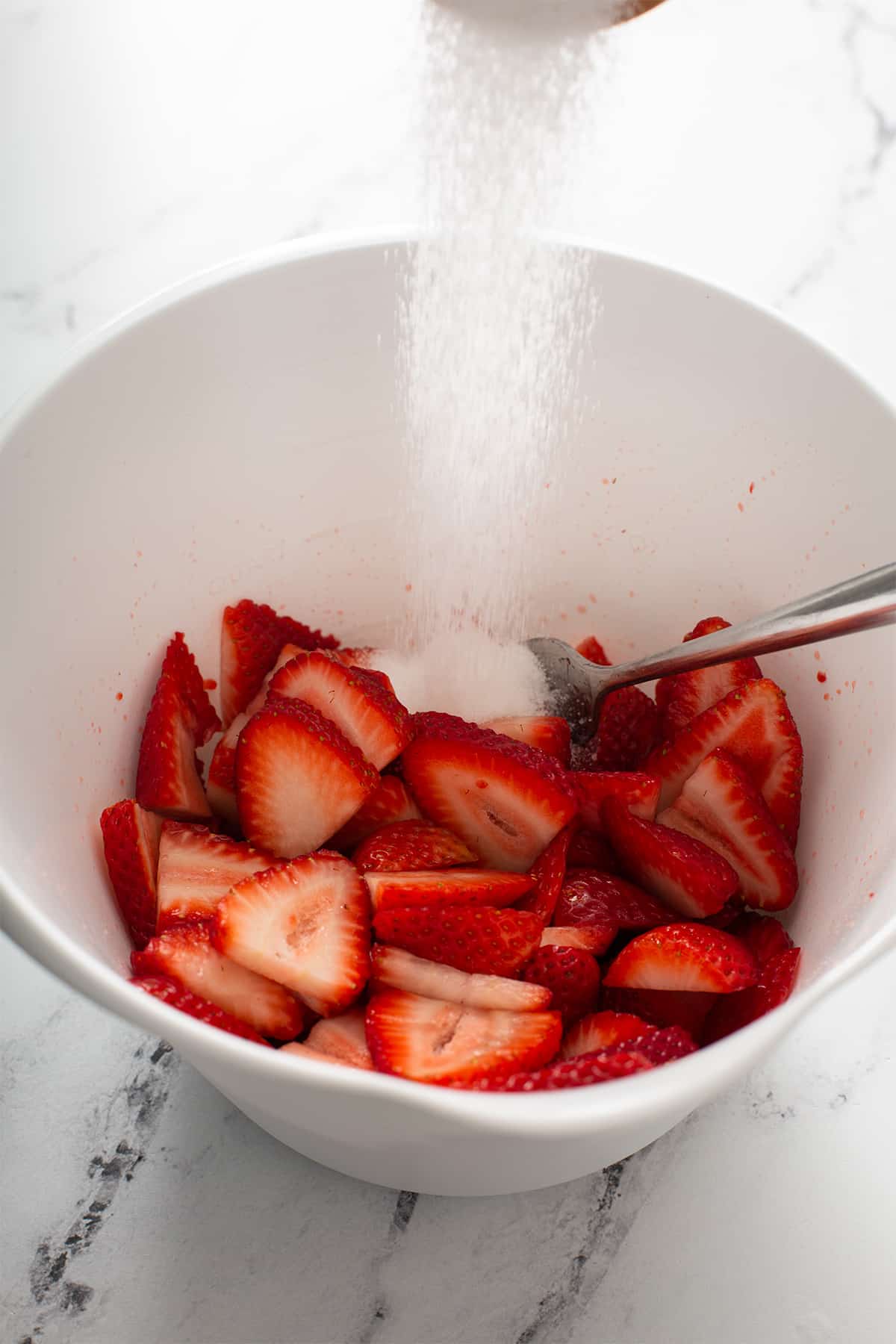Adding sugar into a bowl of fresh strawberries cut up into slices