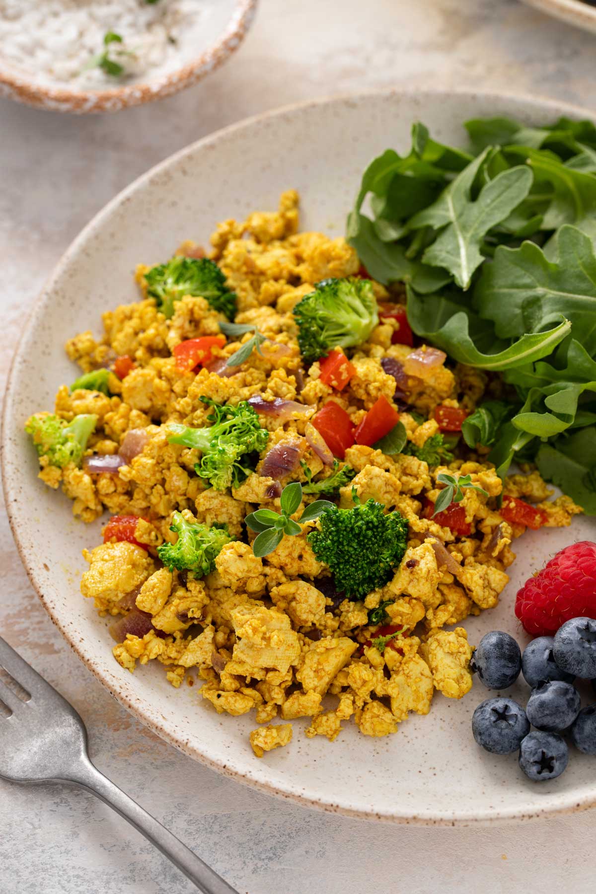 Tofu scramble with optional broccoli, red bell pepper and onion on a plate with arugula and fresh veggies.
