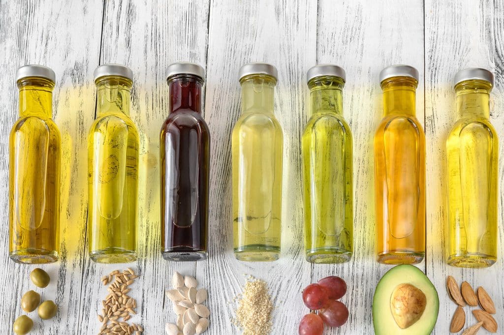 A row of bottled cooking oils, each with its source below it, such as olives, seeds, avocado and almonds.