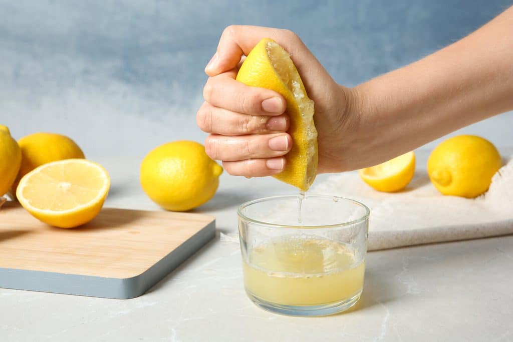 A hand squeezing a lemon half into a small glass of juice, with more lemons on the counter.