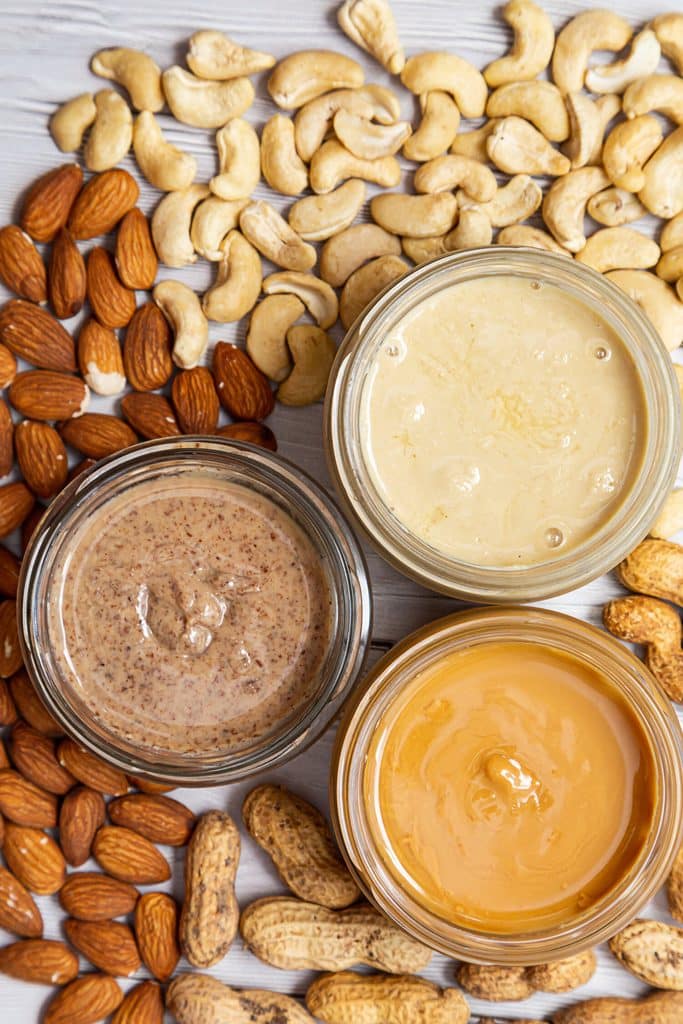 Three jars of nut butter surrounded by cashews, almonds and peanuts.